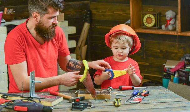Fatherhood concept. Boy, child busy in protective helmet learning to use handsaw with dad. Father, parent with beard teaching little son to sawing while son play with toy saw - Photo, image