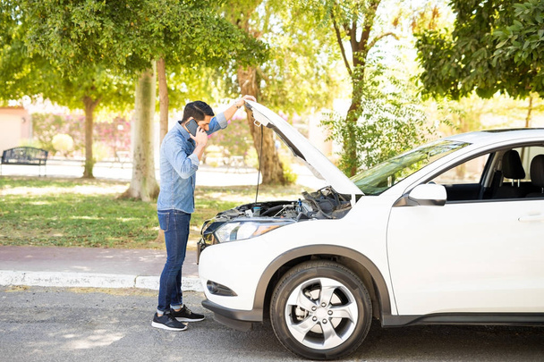 Portrait of young man standing in front of his broken car, looking at car engine and talking on cell phone - Fotografie, Obrázek