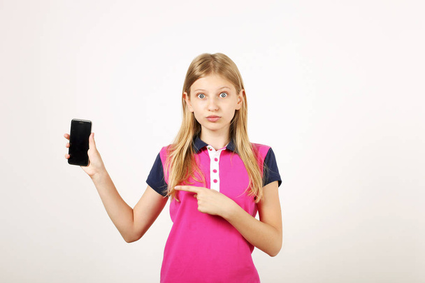 Portrait of casual teen age girl with long natural blond hair, index finger pointing at blank screen cell phone. Unamused young female holding smartphone, showing display. White background, copy space - Fotó, kép