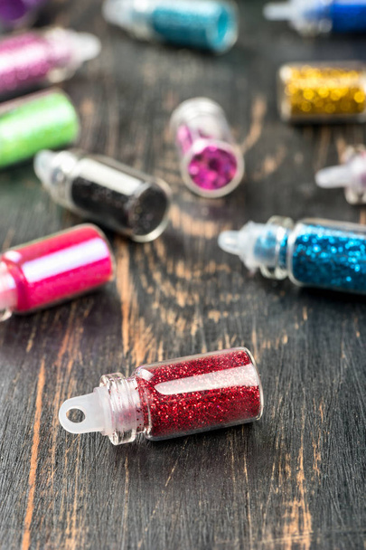 Scattered bottles of colored sparkles for make-up and nail design on wooden background - Фото, изображение