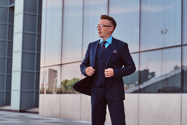 Αυτοπεποίθηση επιχειρηματίας ντυμένοι με ένα κομψό κοστούμι βλέπει μακριά και κουμπί επάνω σακάκι του ενώ στέκεται έξω cityscape φόντο. - Φωτογραφία, εικόνα