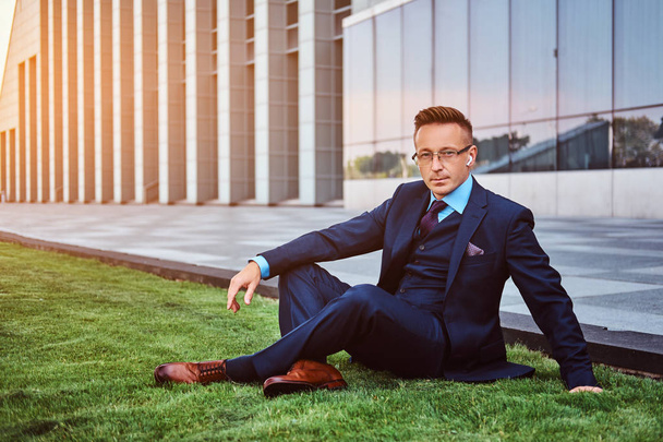 Αυτοπεποίθηση επιχειρηματίας ντυμένος με κομψό κοστούμι που κάθεται σε ένα πράσινο γκαζόν cityscape φόντο.  - Φωτογραφία, εικόνα