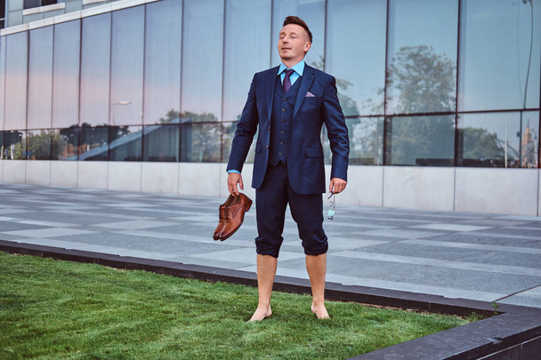 Confident businessman dressed in elegant suit holds his shoes enjoying while standing barefoot on a green lawn against cityscape background.  - Zdjęcie, obraz