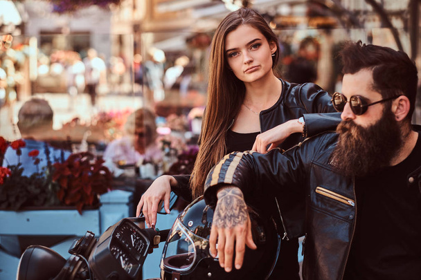 Close-up portrait of a hipster couple - bearded brutal male in sunglasses dressed in a black leather jacket and his young sensual girl standing near, posing against terrace of cafe. - Fotografie, Obrázek