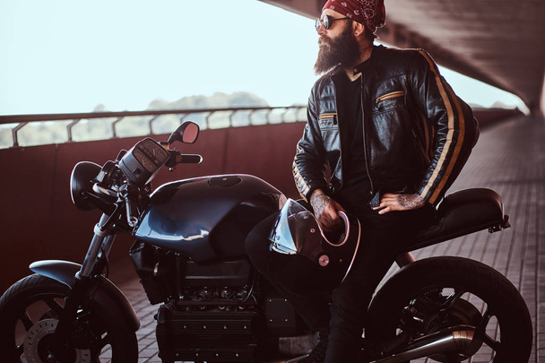 Portréja egy elegáns szakállas férfi öltözött egy fekete bőr kabát és tarka selyemkendő napszemüveg ül a saját rendelésre készült retro motorkerékpár, keres el. - Fotó, kép