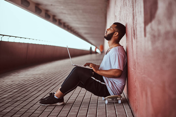 Замислений афро-американський хлопець, одягнений у білий бородатий сорочки й спортивні шорти тримає на ноутбуці сидячи на скейтборді під мостом, спираючись на стіні. - Фото, зображення