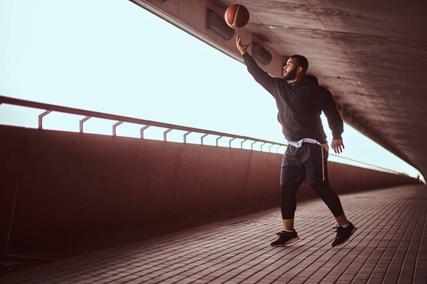 Muotokuva tummaihoinen kaveri pukeutunut musta huppari ja urheilu shortsit pelaa koripalloa kävelyllä sillan alla
. - Valokuva, kuva