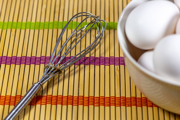 A group of eggs inside a deep white bowl next to a whisk waiting for the chef to use them in a meal on a bamboo mat - Photo, image