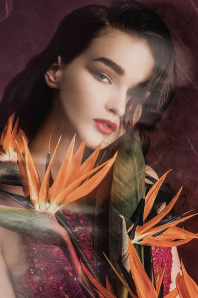 Beautiful girl model in a long red dress on a background of color wine with a bouquet of flowers strelitzia. - Φωτογραφία, εικόνα