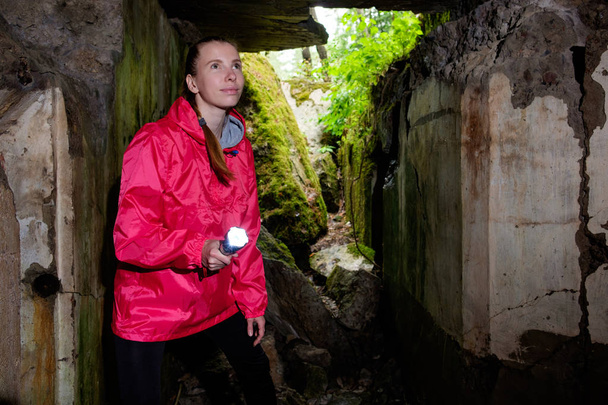 Young woman with lightern, weared in red watercoat, explores ancient fortress cave - Photo, Image
