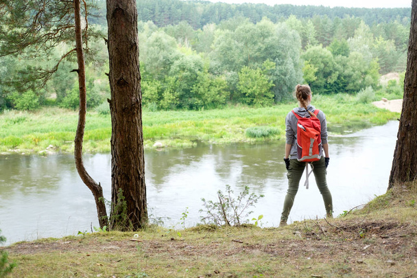 Young redhead woman travels with red travel backpack in summer forest, stands on the bank of river and watches faraway. Concept travel. - Photo, Image