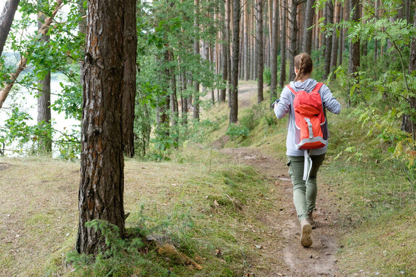 Jonge roodharige lang haar vrouw reist met rode reizen rugzak in zomer bewolkt bos. Concept reizen. - Foto, afbeelding