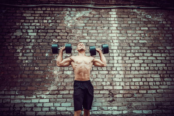 Muscular guy doing exercises with dumbbell against a brick wall - Фото, зображення