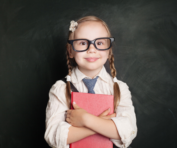 Child with book. Small schoolgirl in uniform clothes holding book and smiling against green chalk board background. Back to school, children learning and education concept - Photo, Image