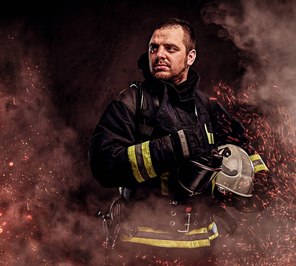 Un bombero profesional vestido con uniforme que sostiene el casco de seguridad en chispas de fuego y humo sobre un fondo oscuro
. - Foto, imagen