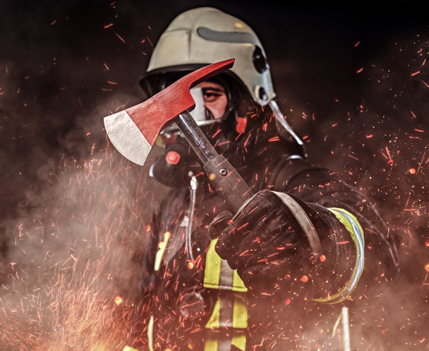 Ein Feuerwehrmann in Uniform und Sauerstoffmaske hält eine rote Axt in Feuerfunken und Rauch über einem dunklen Hintergrund - Foto, Bild