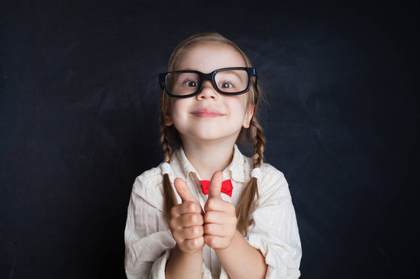 Słodkie dziecko dziewczyna w okularach z kciuk zabawy w klasie na tle pokładzie kredy. Powrót do szkoły, kreatywność i wykształcenie praktyczno - Zdjęcie, obraz