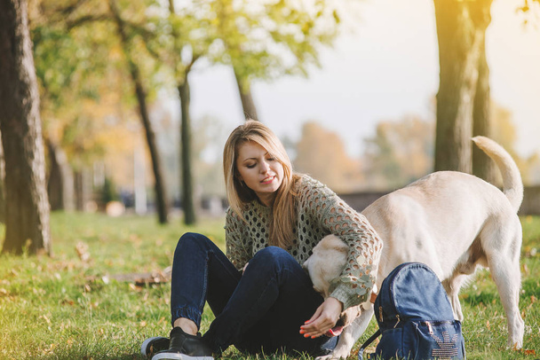 όμορφη ξανθιά κάθεται στο γρασίδι στο πάρκο και παίζει με το σκυλί λαμπραντόρ - Φωτογραφία, εικόνα