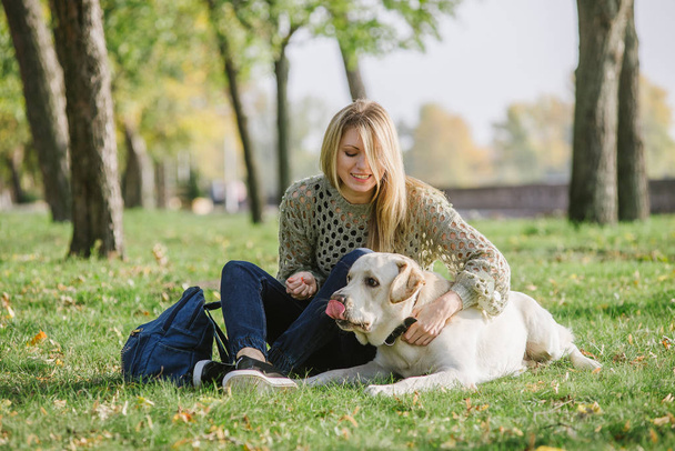 Piękna Blondynka siedzi na trawie w parku i gry z jej pies labrador - Zdjęcie, obraz