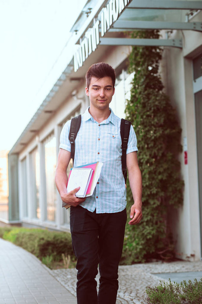 Student holding a notebook and carrying a backpack walking at the front of university building. Young smiling boy wearing blue shirt and dark jeans - Φωτογραφία, εικόνα