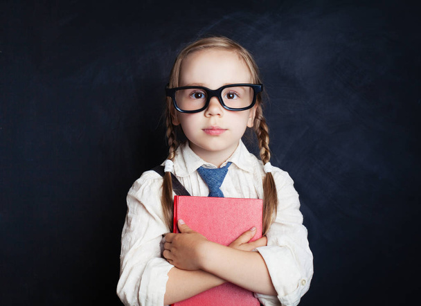 Smart child girl in school uniform clothes with red book on chalk board background. Back to school, happy childhood and elementary school lesson concept. - Φωτογραφία, εικόνα