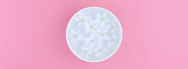 Creative flat lay concept top view of aroma bowl with water and white spring blossoming apple tree flowers on pastel pink paper background with copy space in minimal style, long wide banner - Foto, Imagem