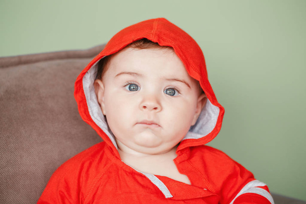 Closeup πορτρέτο του χαριτωμένο αξιολάτρευτο Καυκάσιος έκπληξη μωρό αγόρι με τα μπλε μάτια, φορώντας Μπλούζα φούτερ με κουκούλα κόκκινο sport. Επτά μηνών παιδί που κάθεται στον καναπέ στο σπίτι ψάχνει στην κάμερα - Φωτογραφία, εικόνα