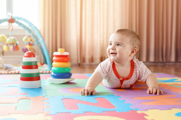 Retrato de lindo adorable niño rubio caucásico sonriente con ojos azules arrastrándose en el suelo en la habitación de los niños niños. Pequeño bebé jugando con juguetes en playmat en casa. Concepto de desarrollo de educación temprana
. - Foto, Imagen