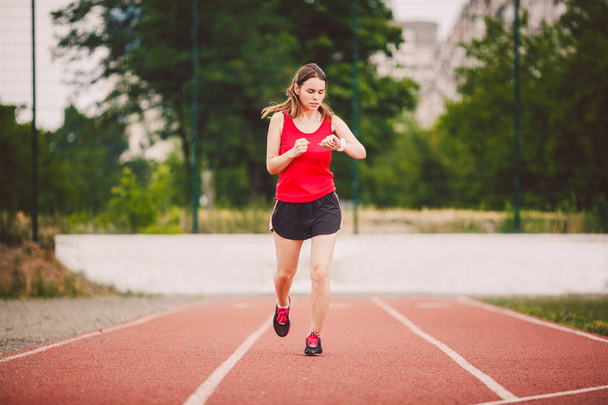 Beautiful young athlete Caucasian woman with big breast in red T-shirt and short shorts jogging, running in the stadium. Hairstyle of pony tail, uses a finger on his hand to press a sports pulseometer - Foto, imagen