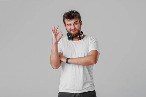 A young man with a beard with wireless headphones around his neck. OK gestures with his fingers, everything is fine. He likes music - Photo, Image