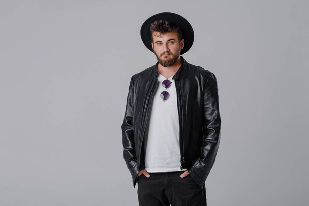 A young cheerful man with a beard in a stylish black leather jacket and hat on a gray background. Smiling at the good news - 写真・画像