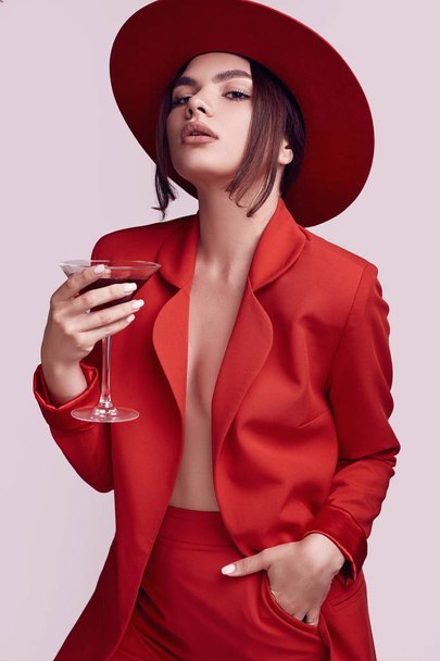 Retrato de mujer hermosa elegante en un traje rojo de moda y sombrero ancho con cóctel de alcohol aislado sobre fondo blanco en el estudio
 - Foto, Imagen