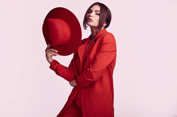 赤のファッショナブルなスーツとスタジオで白い背景に分離された広い帽子でエレガントな美しい女性の肖像画 - 写真・画像