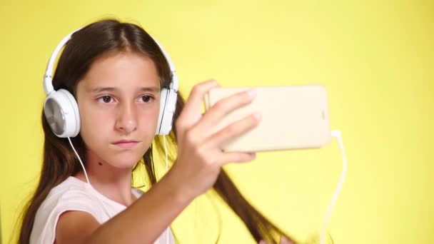 Teenage girl with headphones and mobile phone listening to music on color background. close-up, 4k, slow-motion. makes selfi on smartphone - Materiaali, video