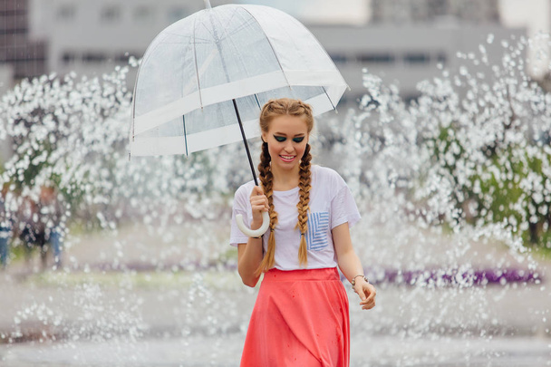 Nuori kaunis tyttö kaksi punokset keltainen saappaat ja läpinäkyvä sateenvarjo seisoo lähellä suihkulähde. Sateinen päivä kaupungissa
. - Valokuva, kuva