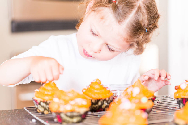Little girl decorating orange icing on cupcakes with sprinkle mix. - Zdjęcie, obraz