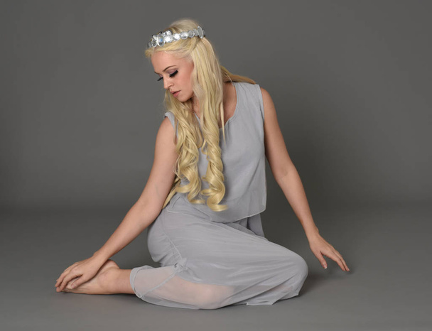 портрет повної довжини блондинки в коронці і блідо-блакитній сукні. сидяча поза на сірому студійному фоні
. - Фото, зображення