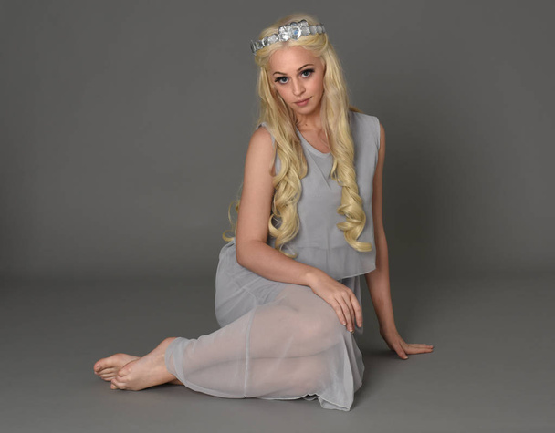 full length portrait of blonde woman wearing crown and pale blue dress. seated pose against a grey studio background. - Foto, Imagen
