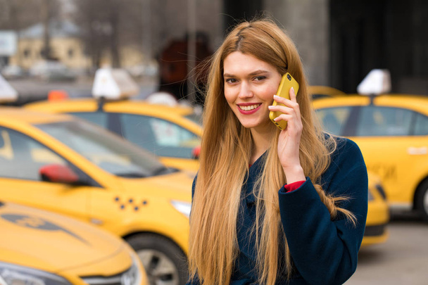 A young girl calls a taxi by phone. Happy blonde woman on smart phone walking in blue coat.  - Φωτογραφία, εικόνα