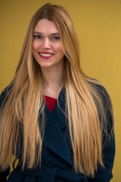 Portrait of a young beautiful woman in blue coat, Happy blonde model against the yellow wall - Foto, imagen