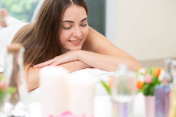 Relaxed woman lying on spa bed for aromatherapy massage in luxury spa with blurred foreground of spa treatment set including aromatic oil, candle and herbal scrub. Wellness and healing concept. - Photo, Image