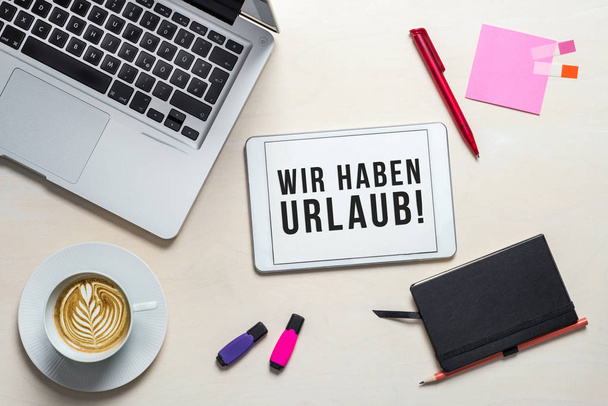 Wir haben Urlaub written in german on tablet meaning Closed for holidays as flatlay from above of an office desk background with notebook - Foto, immagini