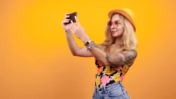 Beautiful gorgeous sexy blonde takes a selfie while wearing a hat, shorts and a swimsuit on yellow orange background. Hot seductive woman - Footage, Video