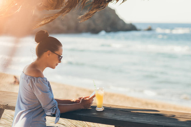 Woman in summer dress and sunglasses making drinking juice and texting messages with smartphone on beach - Foto, Imagen