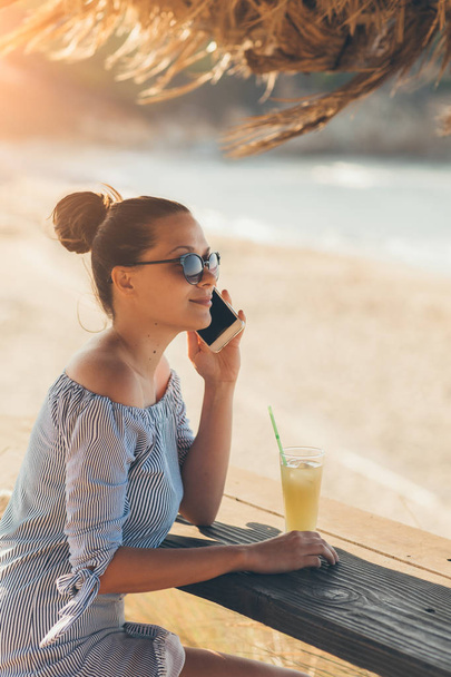 Pretty woman in summer dress drinking cocktail on beach and talking using smartphone - Foto, Imagen