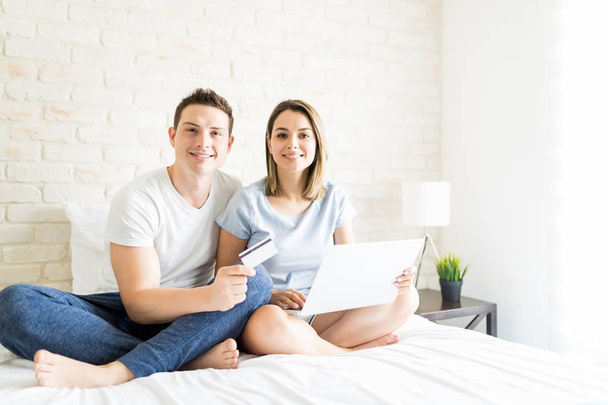Portrait of smiling man and woman shopping for online products using credit card and laptop on bed at home - Zdjęcie, obraz