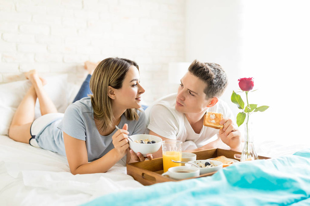 Man and woman talking while having food by breakfast tray in bed at apartment - Photo, Image
