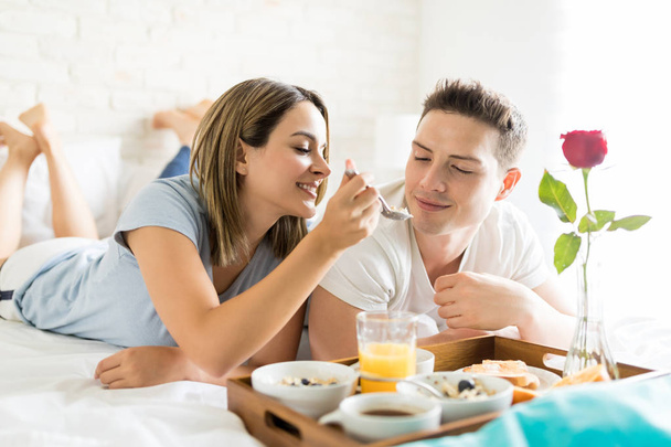 Beautiful young woman feeding boyfriend during breakfast in bed at home - Zdjęcie, obraz