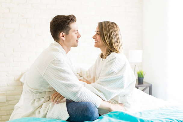 Zijaanzicht van romantische paar badjassen dragen zittend aangezicht tot aangezicht op bed thuis - Foto, afbeelding