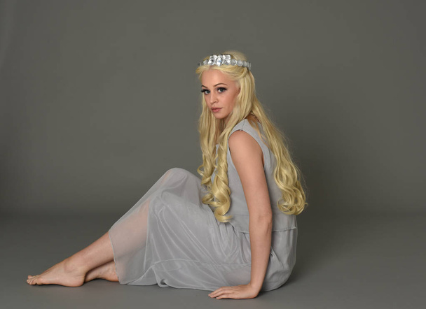 full length portrait of blonde woman wearing crown and pale blue dress. seated pose against a grey studio background. - Photo, Image